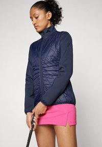 Merimetsävaatteilla varustettu tummansininen vuorattu takki, jossa on pitkät hihat, korkea kaulus ja etuvetoketju; yhdistetty vaaleanpunaiseen hameeseen. Sujuva rakenne ja muodollinen muotoilu.