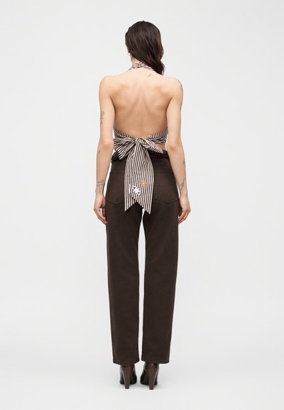 Γυναίκα με μακριά σκούρα μαλλιά που φοράει ένα ριγέ τοπ με τιράντες δεμένο με φιόγκο και σκούρο ψηλόμεσο παντελόνι, στέκεται με την πλάτη προς τα εμπρός.