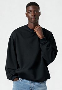 Felpa nera oversize con scollo rotondo, polsini a costine e texture liscia, abbinata a jeans azzurri.