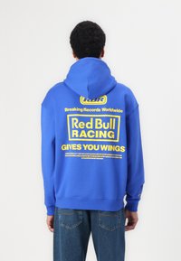 Sudadera azul con capucha, presenta un gran texto gráfico amarillo en la parte posterior: "Red Bull Racing TE DA ALAS" y logotipos adicionales. Diseño y ajuste casual.