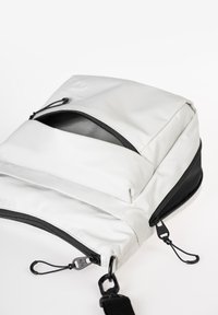 Witte stoffen tas met een gladde textuur, voorzien van een ritsluiting aan de bovenkant en een voorvak, zwarte accenten en verstelbare schouderbandclips.