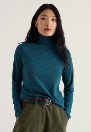 Jeune femme aux longs cheveux foncés portant des boucles d'oreilles en or, un col roulé rayé bleu sarcelle et marine, un pantalon vert olive et une ceinture noire, les mains dans les poches.