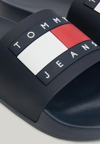 Close-up van een marineblauwe slide-sandaalband met een rechthoekig Tommy Jeans-logo in wit, rood en marineblauwe kleurvlakken en witte letters.