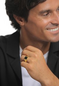 Guld ring med en rektangulær sort sten på en finger, båret af en person iført en sort jakke, der fremhæver glatte, polerede overflader.