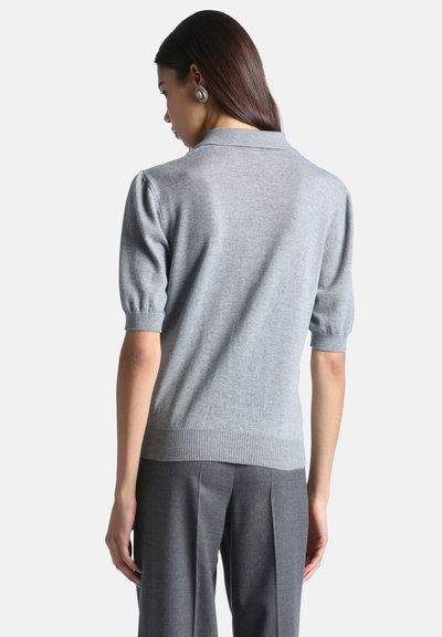 Femme aux longs cheveux foncés portant un pull gris clair à manches courtes et un pantalon gris foncé sur mesure, vue de dos sur fond blanc.