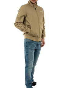 Veste légère beige avec fermeture éclair, col montant, poches latérales et ourlet côtelé. Portée avec un jean bleu et des chaussures noires.