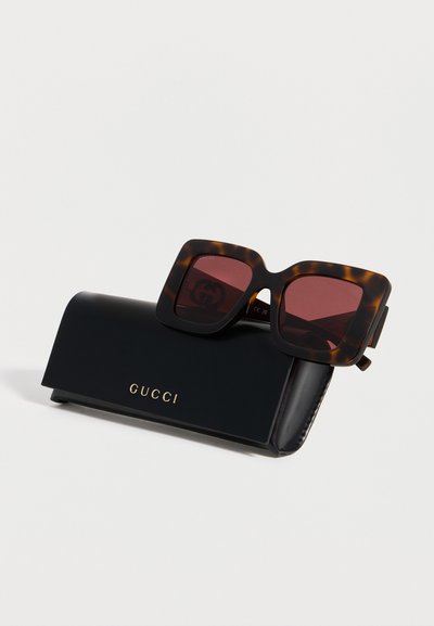 Τετράγωνα γυαλιά ηλίου σχέδιο χελώνας με σκουρόκοκκινα φακούς που ακουμπούν σε μαύρη θήκη Gucci με χρυσό λογότυπο σε λευκό φόντο.