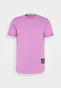 Levandulové tričko s krátkým rukávem a ohrnutými manžetami, s malou černou nášivkou loga Calvin Klein blízko lemu na bílém pozadí.