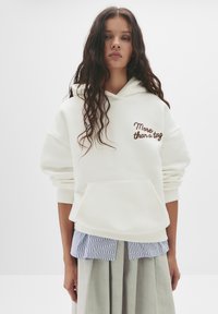 Felpa bianca con una vestibilità rilassata, dotata di una tasca frontale e con "More than a tag" ricamato in marrone. Indossata con una camicia a righe blu e bianca.