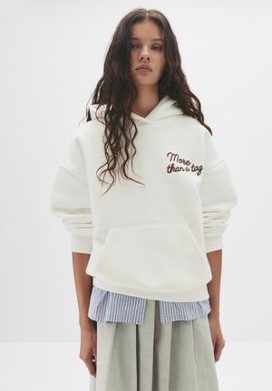 Felpa bianca con una vestibilità rilassata, dotata di una tasca frontale e con "More than a tag" ricamato in marrone. Indossata con una camicia a righe blu e bianca.