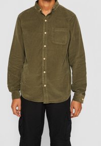 Uomo che indossa una camicia a bottoni in velluto a coste verde oliva con tasca sul petto e pantaloni cargo neri, in piedi contro uno sfondo semplice.