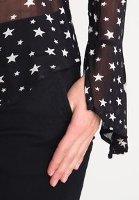 Svart genomskinlig topp med vit stjärntryck; långa ärmar med utställda manschetter. Närbild av ärmdetalj och hand som vilar på svart tyg.