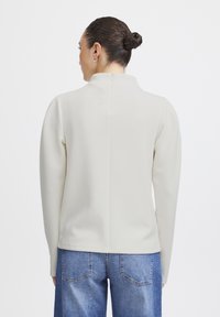 Pull en molleton couleur crème avec un col haut, des manches longues et un design de dos sans couture. Assorti à un jean en denim bleu.