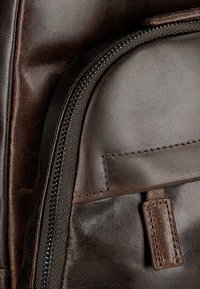 Dettaglio di una borsa in pelle marrone scuro che mostra una superficie liscia, una cerniera e accenti cuciti. Presenta una traccia della cerniera nera a contrasto.