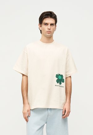 Fiatal férfi visel egy túlméretezett krémszínű pólót, melyen az alsó jobb oldalon egy kis sötétzöld virág és a „marimekko” felirat található, világos farmernadrággal párosítva.