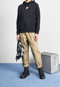 Svart hoodie med en framficka och Nike-logotyp, kombinerad med beiga cargobyxor och svarta kängor. Håller i en svart mönstrad väska.