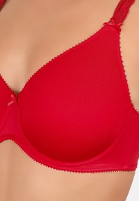 Czerwony biustonosz z miękkiego materiału, w kształcie trójkąta, z regulowanymi koronkowymi ramiączkami i ozdobnymi obszyciami na brzegach. Mały detal w postaci kokardki na środku.