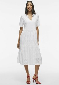 Robe en coton blanche avec un décolleté en V, des manches courtes et des détails en broderie anglaise. La jupe est évasée et se termine sous le genou. Associée à des talons rouges à lanières.