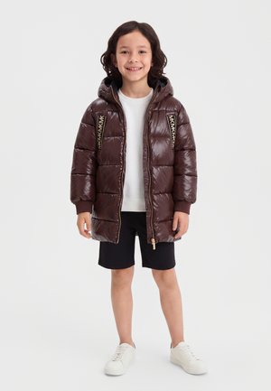 Barn står og smiler, iført en brun dunjakke med hætte, hvid skjorte, sorte shorts og hvide sneakers på en ensfarvet baggrund.