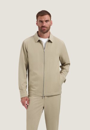 Mand med kort hår og skæg iført beige lynlåstrøje og matchende bukser, stående mod en enkel lys baggrund.