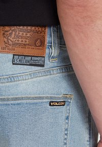 Light blue denim jeans featuring a brown leather patch with an embossed design, black stitching, and a Volcom logo tag on the back pocket.