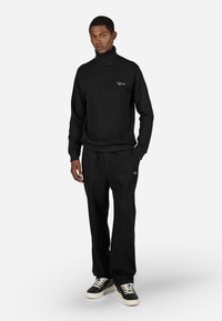 Czarny sweter z golfem o prążkowanej teksturze, w połączeniu z dopasowanymi czarnymi dresami. Zawiera subtelną haftowaną aplikację z logo w białym kolorze.