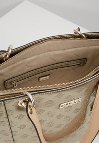 Kabelka ve zlatém odstínu s vembosovaným logem, béžovým interiérem, několika přihrádkami a stříbrnými kovovými doplňky. Ucha na vrchu.