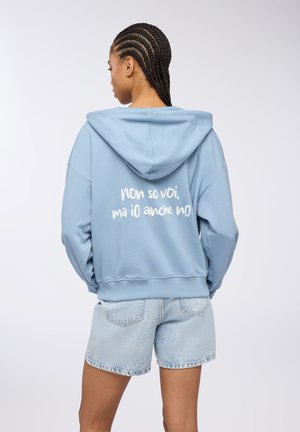 Felpa con cappuccio di un blu chiaro con testo bianco sul retro, abbinata a pantaloni corti di denim chiaro. Il tessuto appare morbido con una vestibilità rilassata.