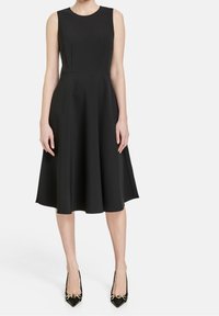 Schwarzes A-Linien-Kleid ohne Ärmel aus einem glatten, matten Stoff, mit rundem Ausschnitt und Falten, kombiniert mit spitzen schwarzen High Heels mit goldenen Akzenten.