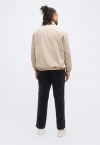 Άνδρας με πλεγμένα μαλλιά στέκεται με την πλάτη προς τα εμπρός φορώντας ένα μπεζ σακάκι, μαύρο παντελόνι και λευκά αθλητικά παπούτσια σε απλό ανοιχτό φόντο.