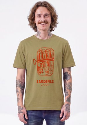 Junger Mann mit lockigem Haar und Tattoos lächelt, trägt ein olivgrünes T-Shirt mit roter Sardinendosen-Grafik und dem Text "SARDINES Portugal".