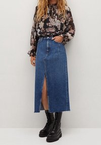 Jupe en denim bleu avec une fente avant, ourlet brut, taille haute et deux poches. Associée à un blouse florale noire et des bottes de combat noires.