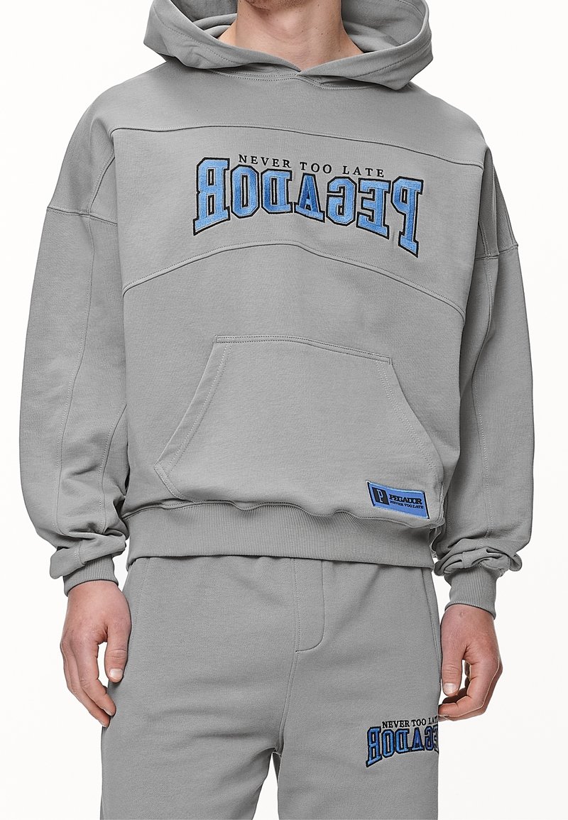 Grå hoodietröja med känguruficka, prydd med blå broderad text "ALDRIG FÖR SENT" och "PECADOR." Ribbad mudd och nederkant. Mjuk tygtextur.