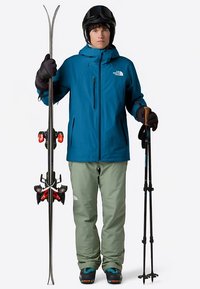 Blue waterproof jacket, green ski pants, black helmet, gloves, holding skis with red bindings and ski poles. Snow-ready outfit for skiing.