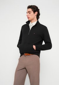 Μαύρο φούτερ με φερμουάρ, με τσέπη καγκουρό και έμβλημα λογότυπου, σε συνδυασμό με ανοιχτό καφέ παντελόνι και ριγέ πουκάμισο με γιακά από κάτω.