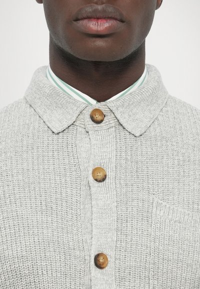 Šviesiai pilkas megztinis su megztu apykakle, puoštas trimis mediniais mygtukais ir krūtinės kišene. Po juo žaliai baltomis juostelėmis dryžuota marškinėliai.