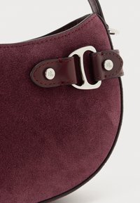 Γωνία τσά�ντας από σούετ σε βαθύ μπορντό με δερμάτινο λουρί και ασημένια πόρπη που φέρει χαραγμένο το λογότυπο "RL", λεπτομερής ραφή στις άκρες του λουριού.