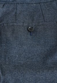 Denim fabric with a dark blue hue, featuring a single black button on a flap pocket, textured surface, and subtle stitching details.