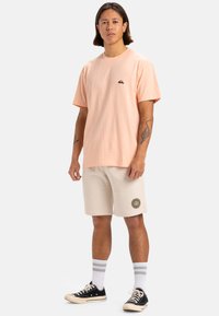 Camiseta de algodón de manga corta en color melocotón con un pequeño logo negro, combinada con pantalones cortos beige que tienen un parche bordado, zapatillas negras y calcetines a rayas.