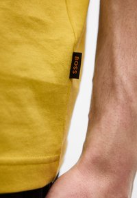 Gros plan sur l'ourlet d'un vêtement jaune avec une étiquette de marque noire "BOSS" et un avant-bras poilu à côté.