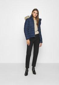 Chaqueta acolchada azul marino con capucha forrada de pelo, diseño de costuras verticales, combinada con un suéter beige y pantalones negros, complementada por botines negros.