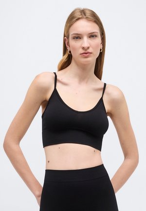 Ung kvinde med langt, blondt hår iført gyldne hoop-øreringe og en sort cropped top med spaghetti-stropper foran en ensartet lys baggrund.