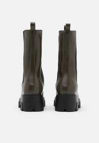 Bottines en synthétique marron avec une finition brillante, des panneaux élastiques sur les côtés et des semelles en caoutchouc noir épaisses. Présente des tirettes au niveau des talons.