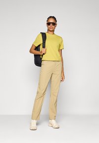 Maglietta gialla a maniche corte abbinata a pantaloni beige chiaro. L'outfit è completato da uno zaino nero e occhiali da sole oversize.