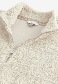 Pull-over confortable en fausse fourrure shearling crème avec un col haut, doté d'une fermeture éclair argentée et d'une texture douce et duveteuse. Étiquette de taille visible à l'intérieur.