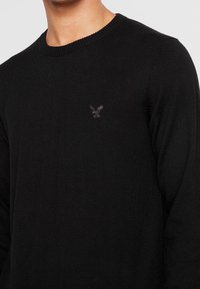Black knitted sweater with a round neck. Features a subtle embroidered eagle logo on the chest. Soft texture and fitted silhouette.