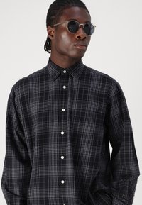 Camisa de cuadros negra y gris con cuello clásico, mangas largas y botones blancos. Modelo con gafas de sol redondas y oscuras.