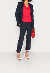 Completo blu navy con una camicetta rossa a bottone, caratterizzata da un colletto arricciato. Sandali con tacco bianchi e una borsa rossa completano l'outfit.