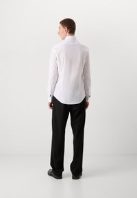 Weißes Langarmhemd mit taillierter Passform, klassischem Kragen und Knopfmanschetten, kombiniert mit schwarzen Hosen und schwarzen Schuhen.
