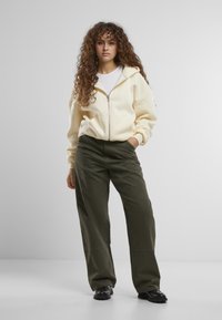 Helle gelbe Zip-Hoodie über einem weißen Shirt, kombiniert mit dunkelgrünen weiten Hosen. Hat lockige Haare und schwarze Schuhe.